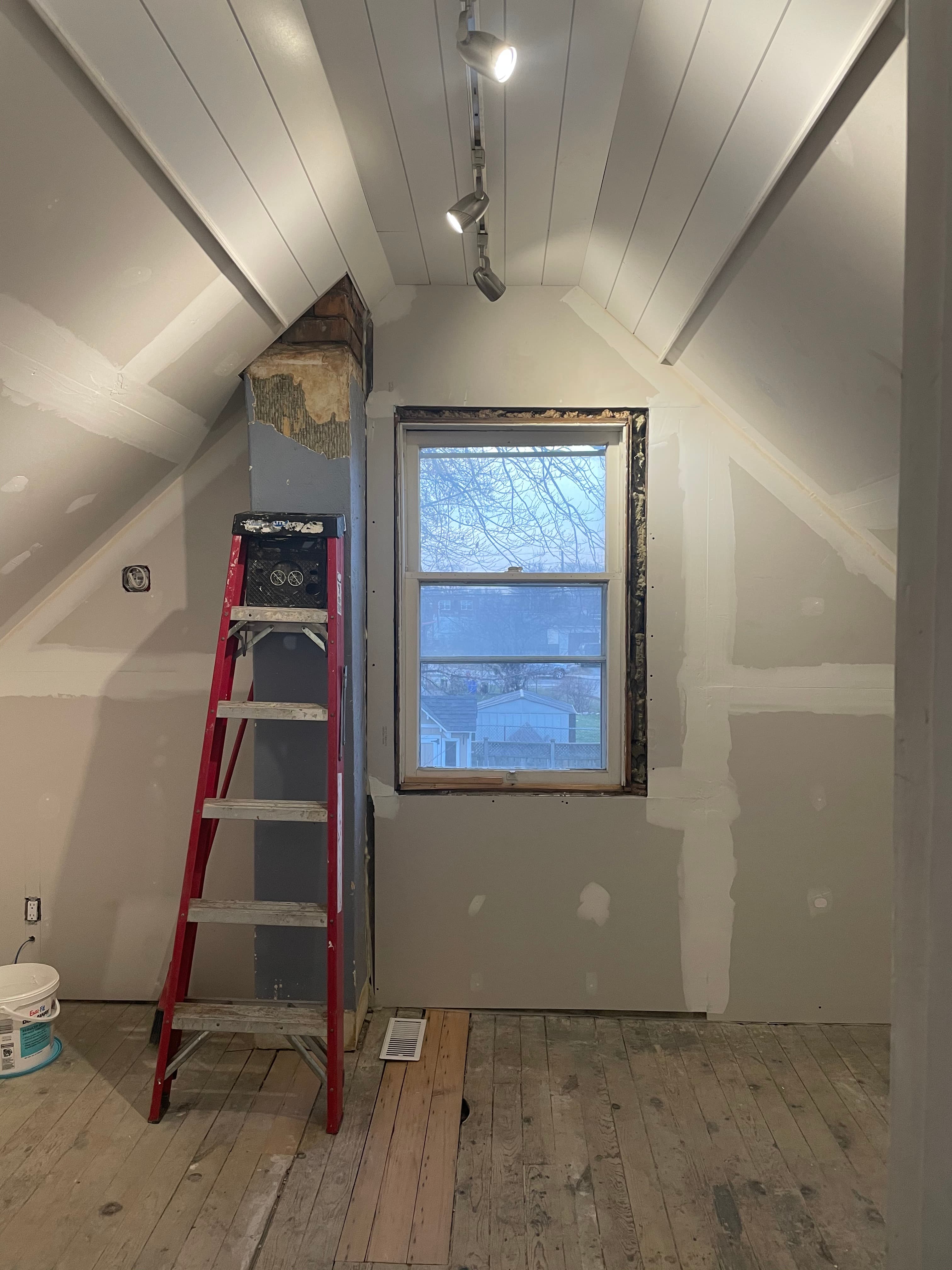Attic room under renovation with drywall work and a ladder beside a window opening.
